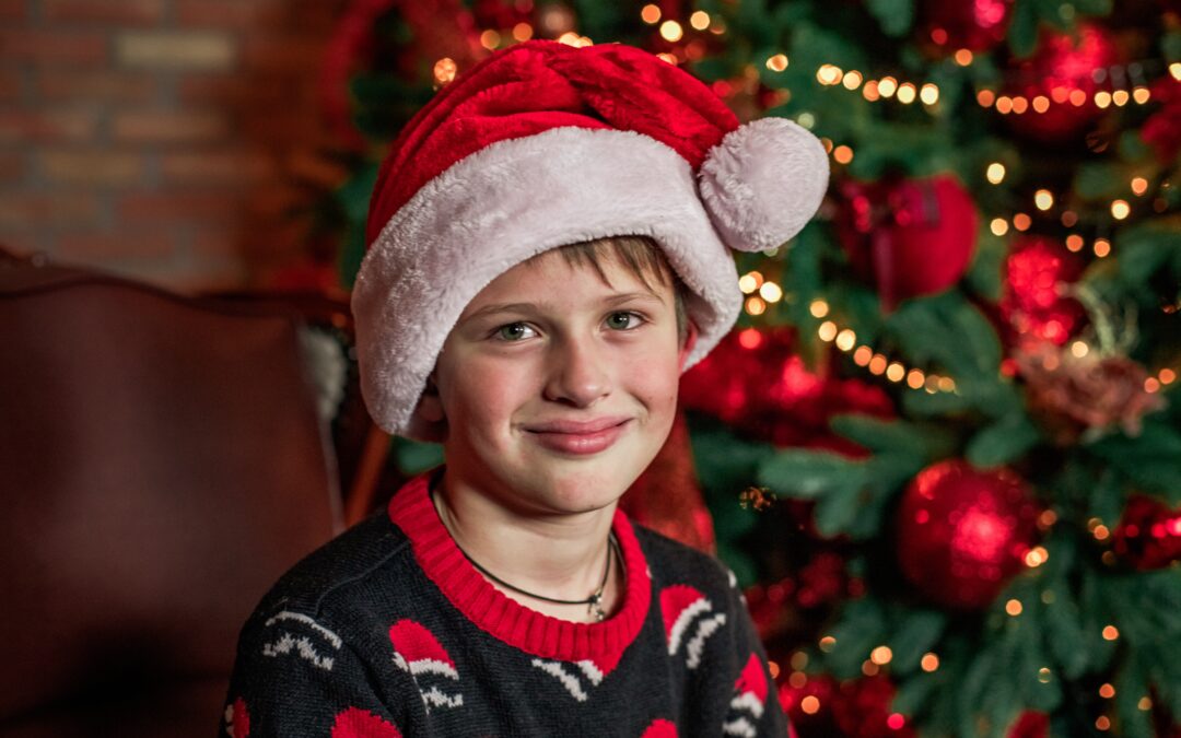 Young child wearing Santa hat and Christmas sweater smiling in front of decorated tree.